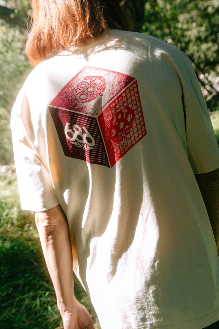 A person is standing outdoors in sunlight with greenery in the background, wearing the 686 Men's Unit Relaxed SS Tee by 686—a light-colored 100% cotton men’s t-shirt featuring a red and white geometric cube graphic with the number 69 on the back.