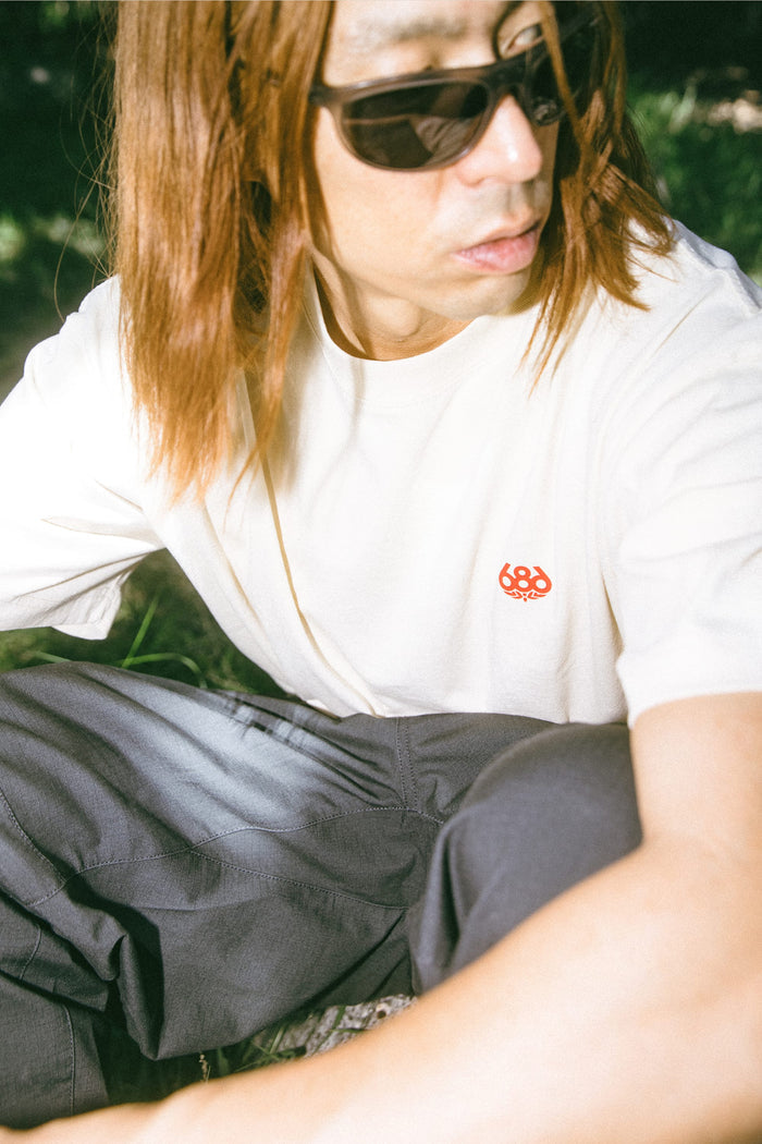 A person with long brown hair, wearing sunglasses and a 686 Men's Unit Relaxed SS Tee (a cream 100% cotton t-shirt with a red logo), paired with dark pants, sits outdoors in sunlight, looking to the side.