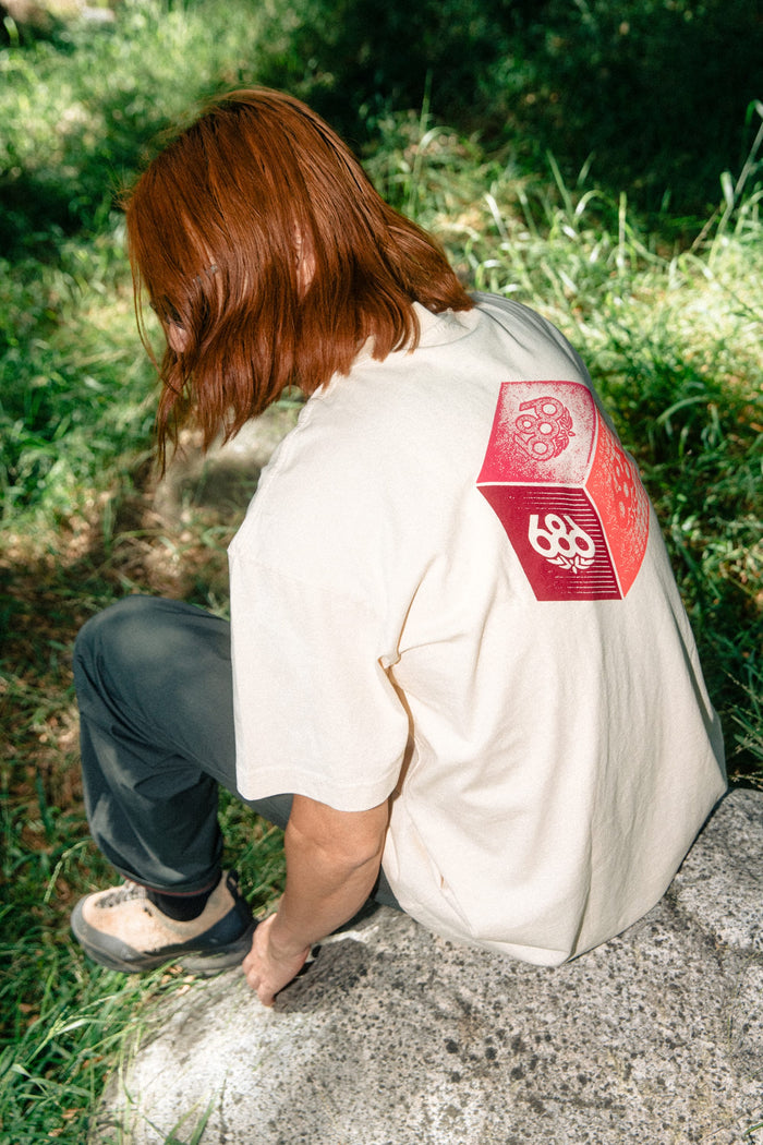A person with long red hair sits on a rock outdoors, wearing the 686 Men's Unit Relaxed SS Tee—a cream 100% cotton t-shirt by 686 featuring a red geometric design on the back. The shirt stands out against the grassy, sunlit setting.