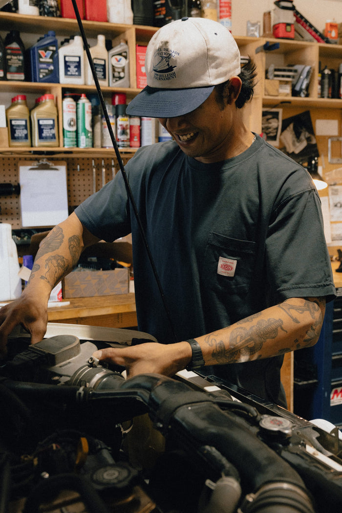A man wearing a cap and a 686 Men's Unwork Authentic SS Pocket Tee smiles while working on an engine in a cluttered workshop, with shelves of bottles and tools visible in the background.