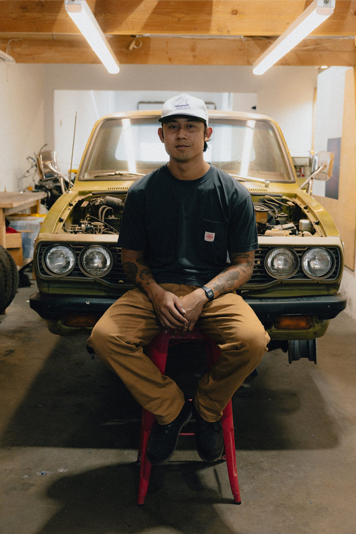 A man wearing a cap, tan pants, and the 686 Men's Unwork Authentic SS Pocket Tee by 686 sits on a stool in a garage, positioned in front of a vintage yellow car with its hood open.