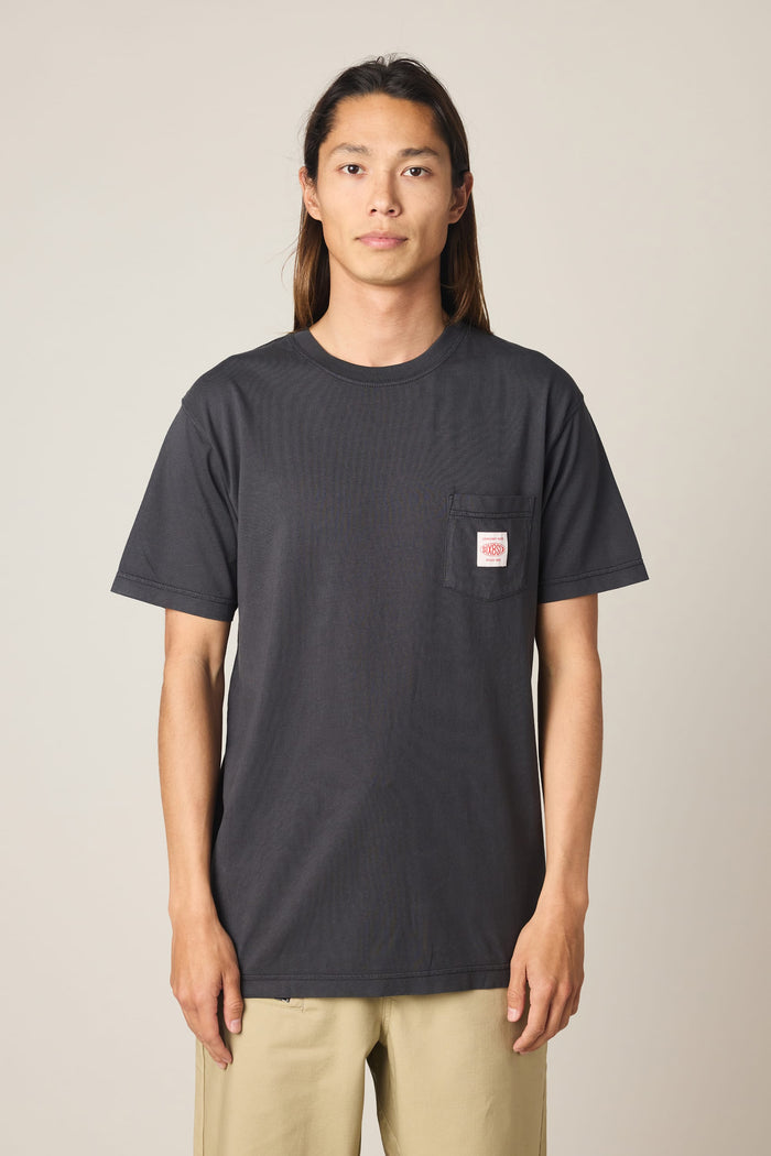 A person with long brown hair wears the 686 Men's Unwork Authentic SS Pocket Tee by 686, a black 100% cotton men's t-shirt featuring a small white and red logo on the chest pocket, paired with beige pants. They stand against a plain light background, facing the camera.