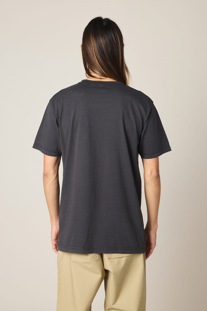 A person with long brown hair stands facing away, wearing the 686 Men's Unwork Authentic SS Pocket Tee by 686 in plain dark gray 100% cotton, paired with light beige pants against a neutral background.