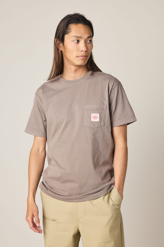 A person with long hair wears the 686 Men's Unwork Authentic SS Pocket Tee in taupe—crafted from 100% cotton and detailed with a small white and red patch on the chest pocket—styled with light beige pants and standing against a plain light background.
