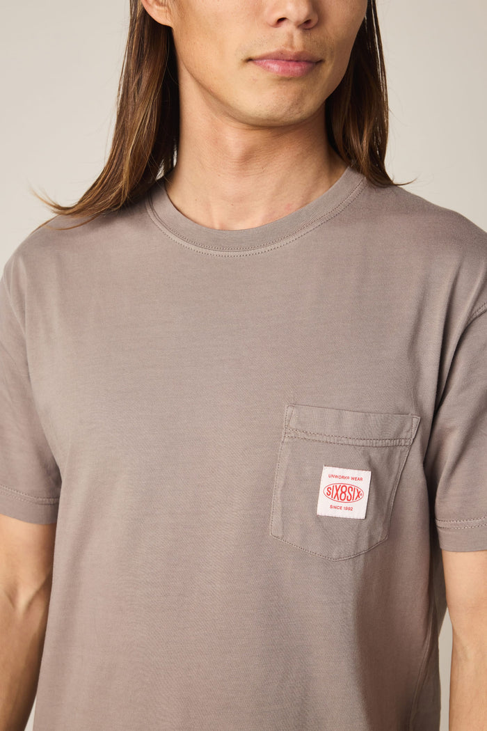 A person with long brown hair wears the 686 Men's Unwork Authentic SS Pocket Tee, a taupe 100% cotton t-shirt from 686 featuring a chest pocket. The pocket displays a red and white SixSox label with smaller text above it. The person's face is partially visible.