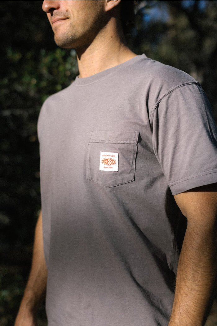 A man is wearing the 686 Men's Unwork Authentic SS Pocket Tee in light gray, made from 100% cotton, featuring a chest pocket with a small rectangular patch. The background is blurred greenery, with sunlight highlighting parts of his shirt and face.