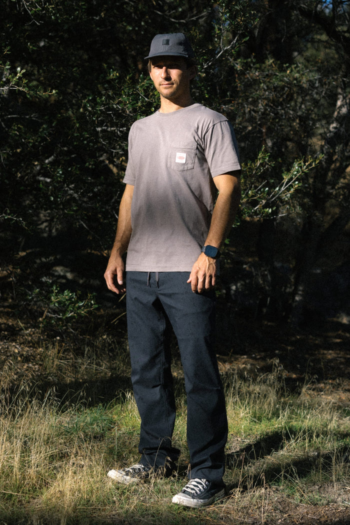 A person wearing a 686 Men's Unwork Authentic SS Pocket Tee in gray, paired with black pants, a black cap, and sneakers, stands outdoors on grass with trees and sunlight in the background.
