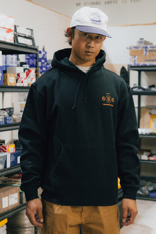 A person wearing a white cap, brown pants, and the 686 Men's Unwork Spec Athletic LS Hoody by 686 stands in a room filled with shelves of boxes and parts, looking directly at the camera.