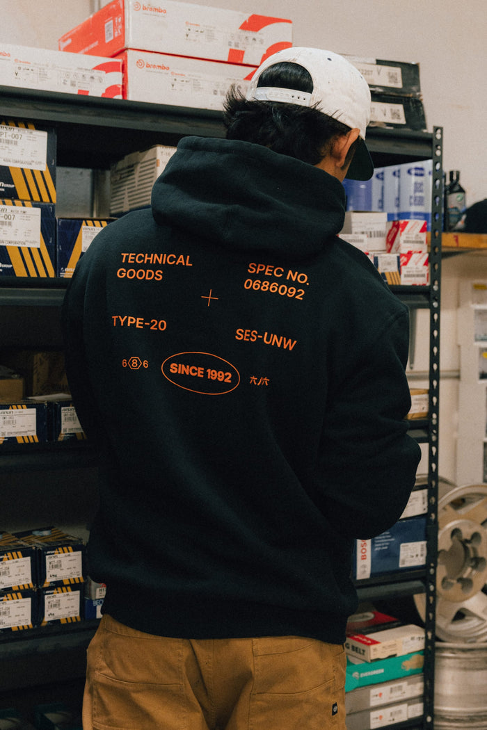 A person wearing the 686 Men's Unwork Spec Athletic LS Hoody stands with their back to the camera, displaying its black fabric and orange technical text, in front of shelves filled with various boxes and supplies.