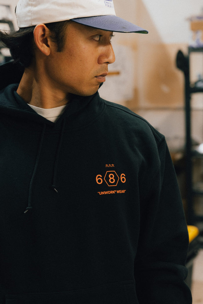 A person wearing the 686 Men's Unwork Spec Athletic LS Hoody in black, featuring orange "686 UNWORK WEAR" text, and a white cap looks to the side in an indoor setting.