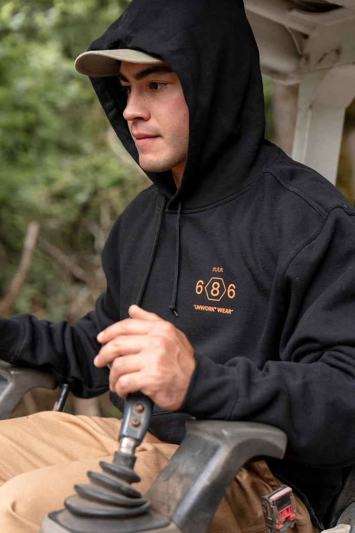 Wearing the 686 Men's Unwork Spec Athletic LS Hoody in black, a person pairs it with tan pants while operating outdoor machinery using a joystick control. Trees and greenery are visible in the blurred background.