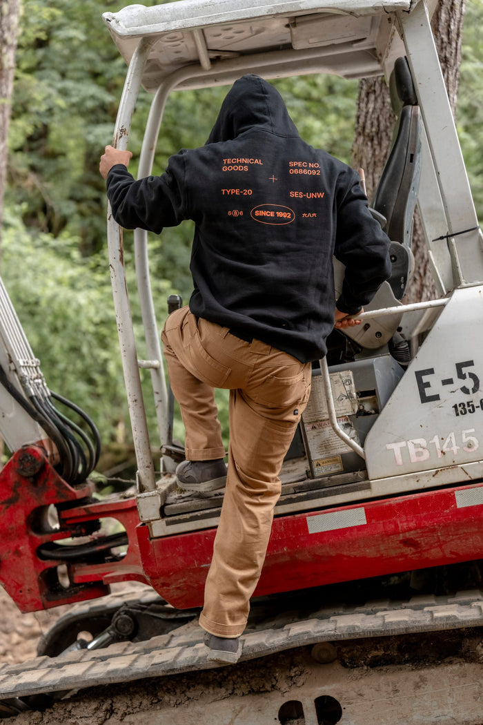Wearing the 686 Men's Unwork Spec Athletic LS Hoody, a person in tan pants climbs into a white and red excavator in a wooded outdoor setting.