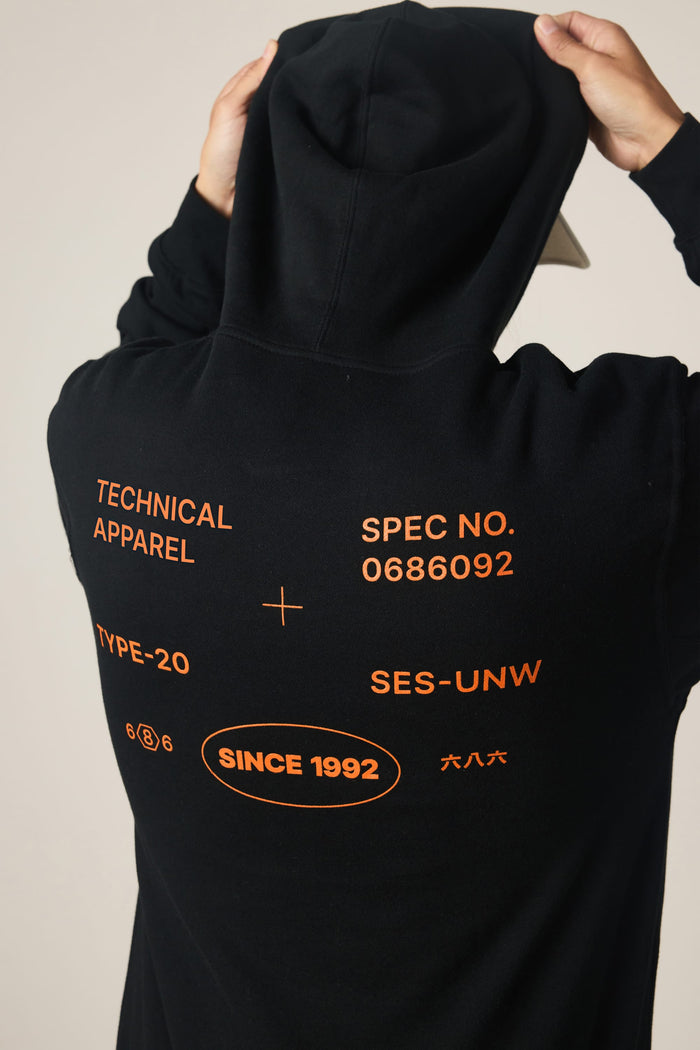 A person wears the 686 Men's Unwork Spec Athletic LS Hoody—a black cotton sweatshirt by 686—featuring bold orange text on the back that reads "TECHNICAL APPAREL, SPEC NO. 0686092, TYPE-20, SES-UNW, and SINCE 1992." The individual holds the hood with both hands.