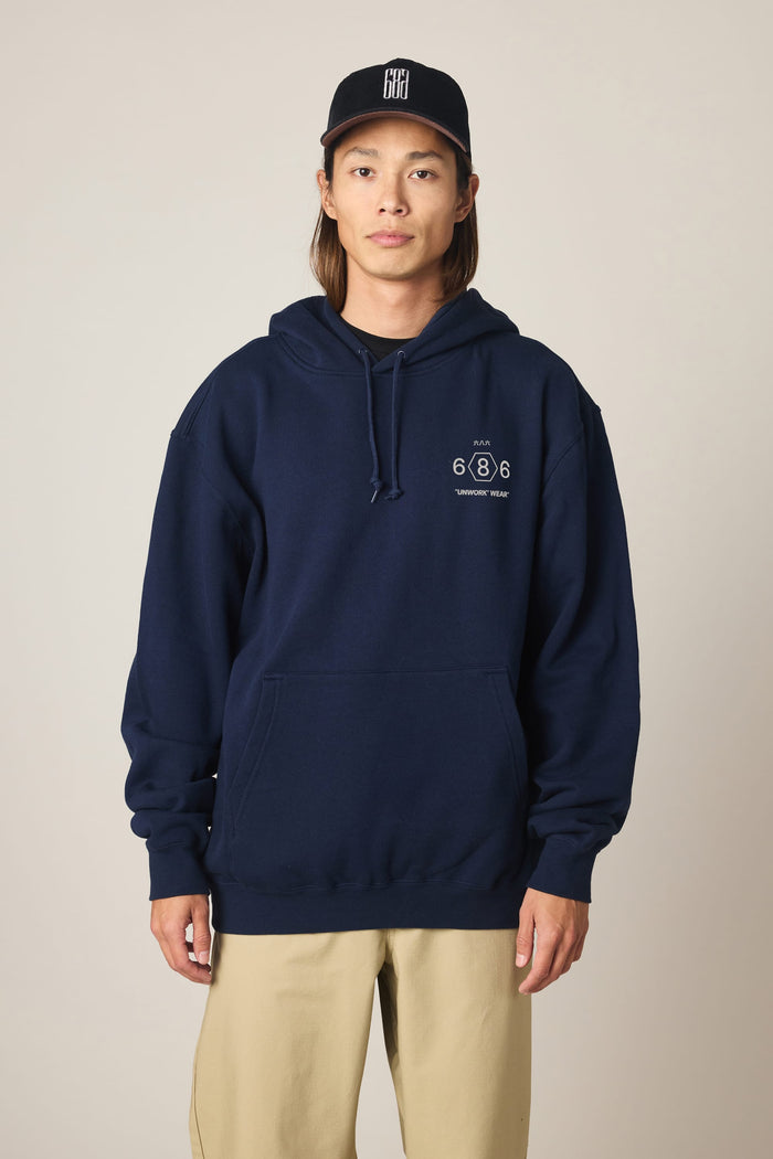 A person wearing the 686 Men's Unwork Spec Athletic LS Hoody in navy, featuring the 686 logo and text on the left chest, paired with beige pants and a black baseball cap, stands against a plain beige background.