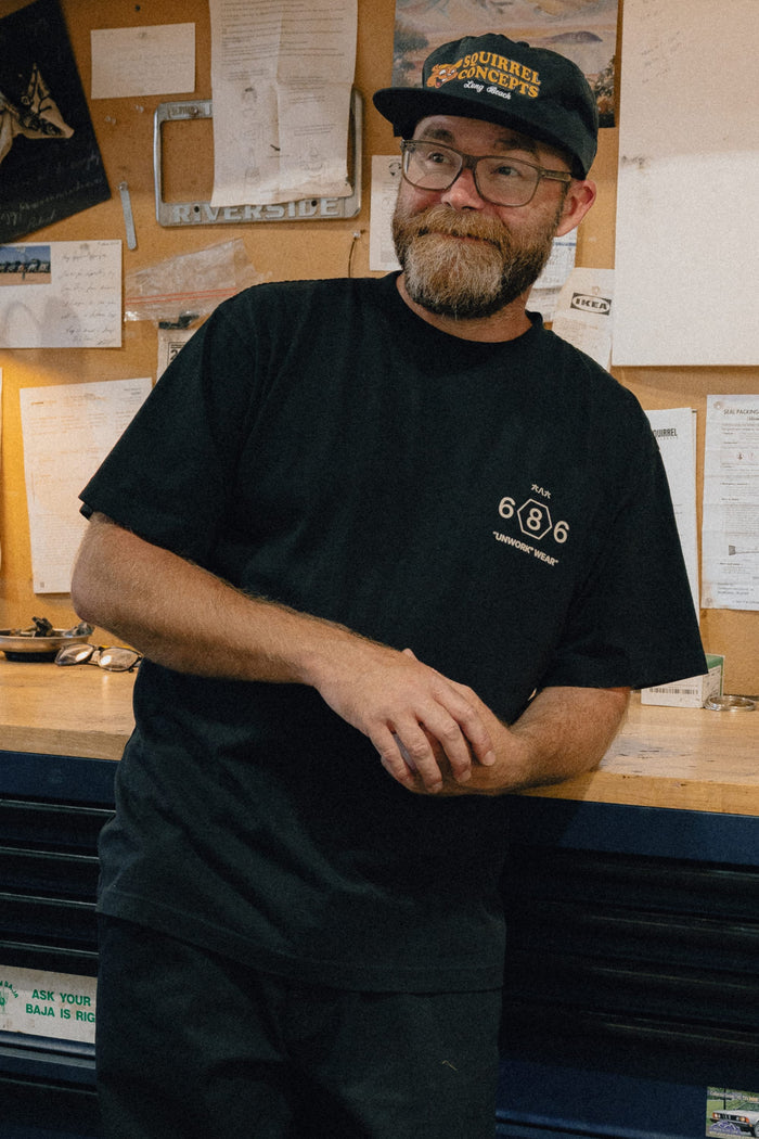A man with glasses and a beard, wearing the 686 Men's Unwork Spec Relaxed SS Tee and a cap, stands leaning against a workbench in front of a bulletin board with papers and tools.