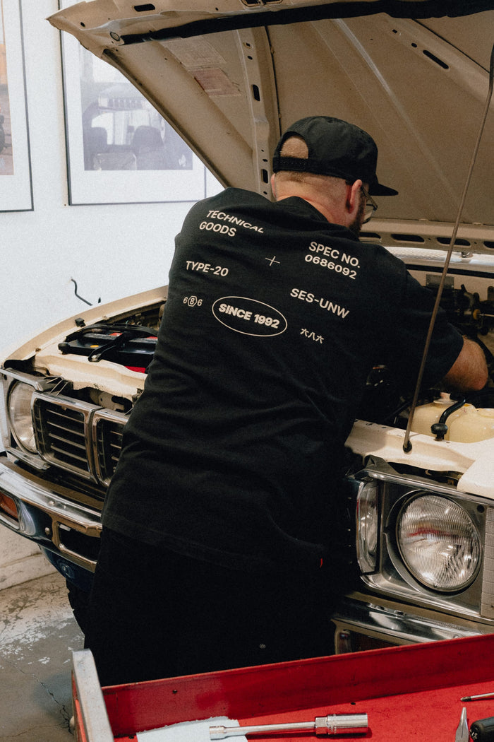 A person wearing a 686 Men's Unwork Spec Relaxed SS Tee in black works under the open hood of a vintage white car inside a garage.