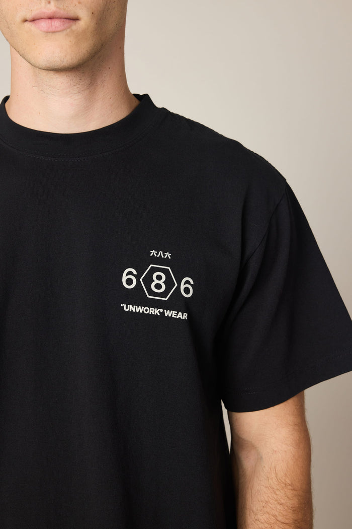A person models the 686 Men's Unwork Spec Relaxed SS Tee, a 100% cotton black t-shirt by 686, featuring “UNWORK WEAR” and Japanese lettering in white on the chest along with a hexagon emblem. The image captures them from just below the face to the upper torso.