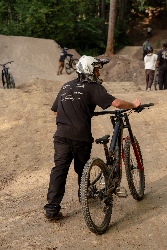 A person dressed in black clothing and a white helmet, wearing the 686 Men's Unwork Spec Relaxed SS Tee by 686, pushes a mountain bike up a dirt trail in a wooded area, with other cyclists and bikes visible in the background.