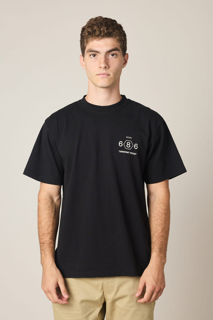 A young man with short curly hair wears the 686 Men's Unwork Spec Relaxed SS Tee—a loose, black 100% cotton t-shirt featuring a small “686” logo on the left chest—paired with tan pants. He stands facing forward against a plain, light background, showcasing the comfort and breathability of this classic piece from 686.