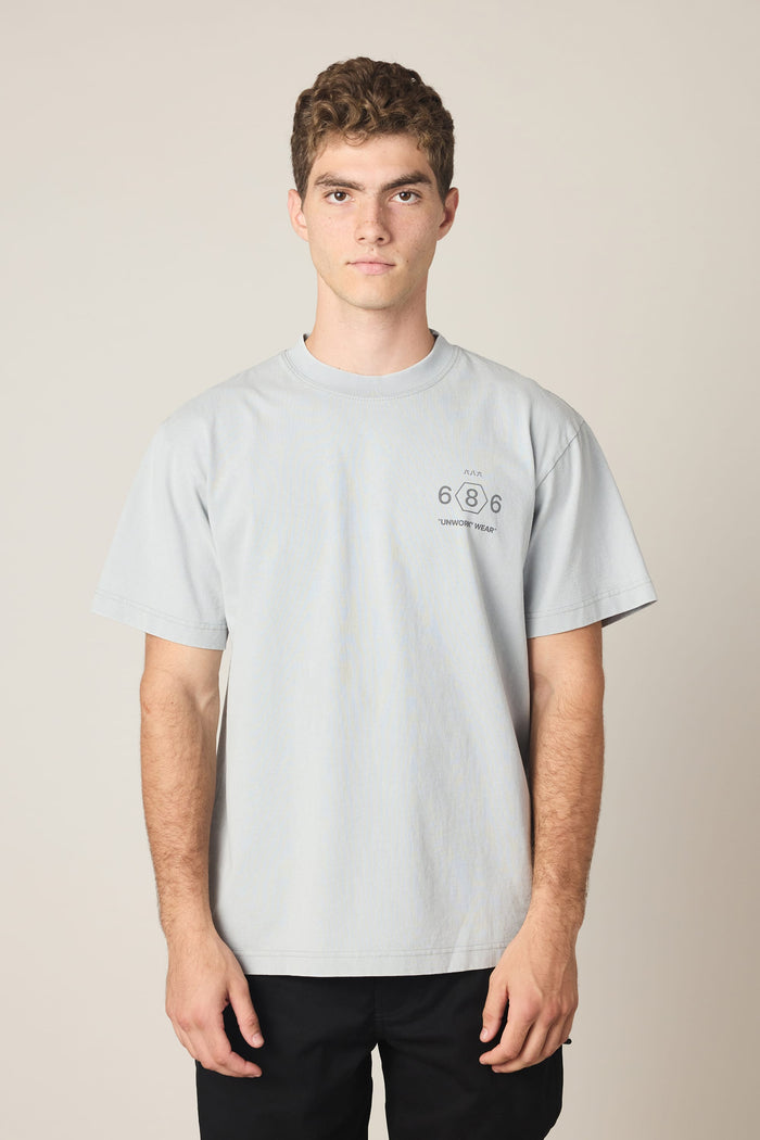 A young man with short curly hair wears the 686 Men's Unwork Spec Relaxed SS Tee, a light gray 100% cotton t-shirt from 686 featuring "686" and small text on the left chest. He stands facing forward against a plain beige background with a neutral expression.