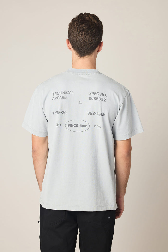 A person is seen from behind wearing the 686 Men's Unwork Spec Relaxed SS Tee, a light gray cotton t-shirt by 686. The back features various technical-style text and symbols such as "TECHNICAL APPAREL," "SINCE 1992," and other numbers and codes, giving it a unique graphic design that blends comfort with modern style.