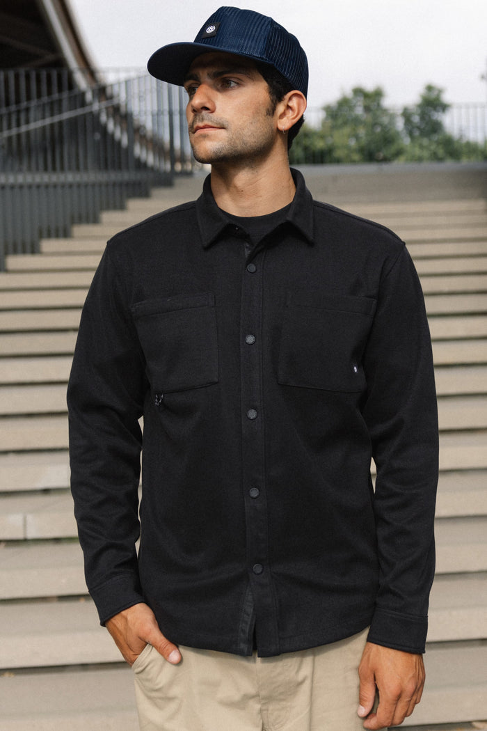 A man wearing the 686 Men's Whisper Knit™ Flannel and a navy cap stands outdoors in front of concrete stairs, looking to the side with one hand in his pocket.