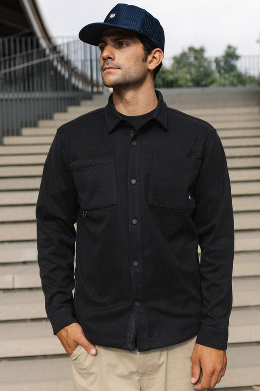 A man wearing the 686 Men's Whisper Knit™ Flannel and a navy cap stands outdoors in front of concrete stairs, looking to the side with one hand in his pocket.
