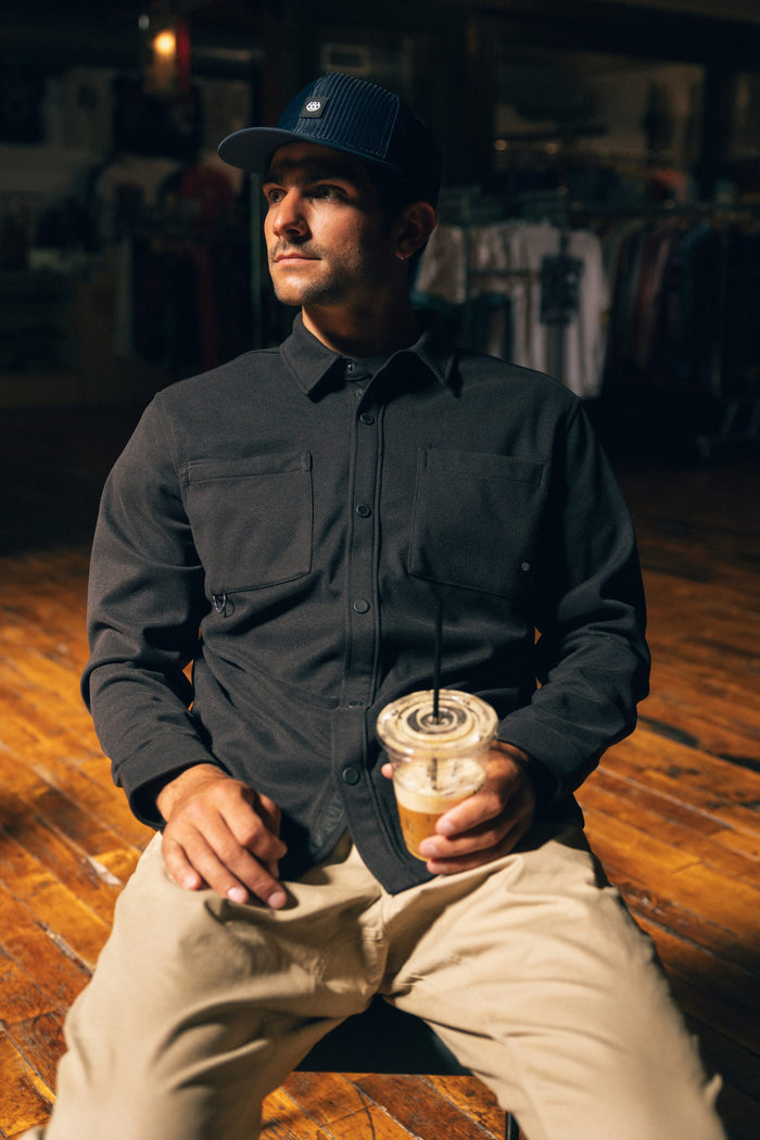 A man wearing the 686 Men's Whisper Knit™ Flannel by 686 and light pants sits on a wooden floor, holding an iced coffee with a straw. He gazes off to the side in a dark cap, while blurred clothing racks appear in the background.