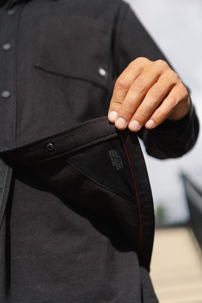 A person’s hand holds open the front of the 686 Men's Whisper Knit™ Flannel, revealing the inside tag and red stitching detail on this black 100% polyester shirt by 686. The background is blurred.