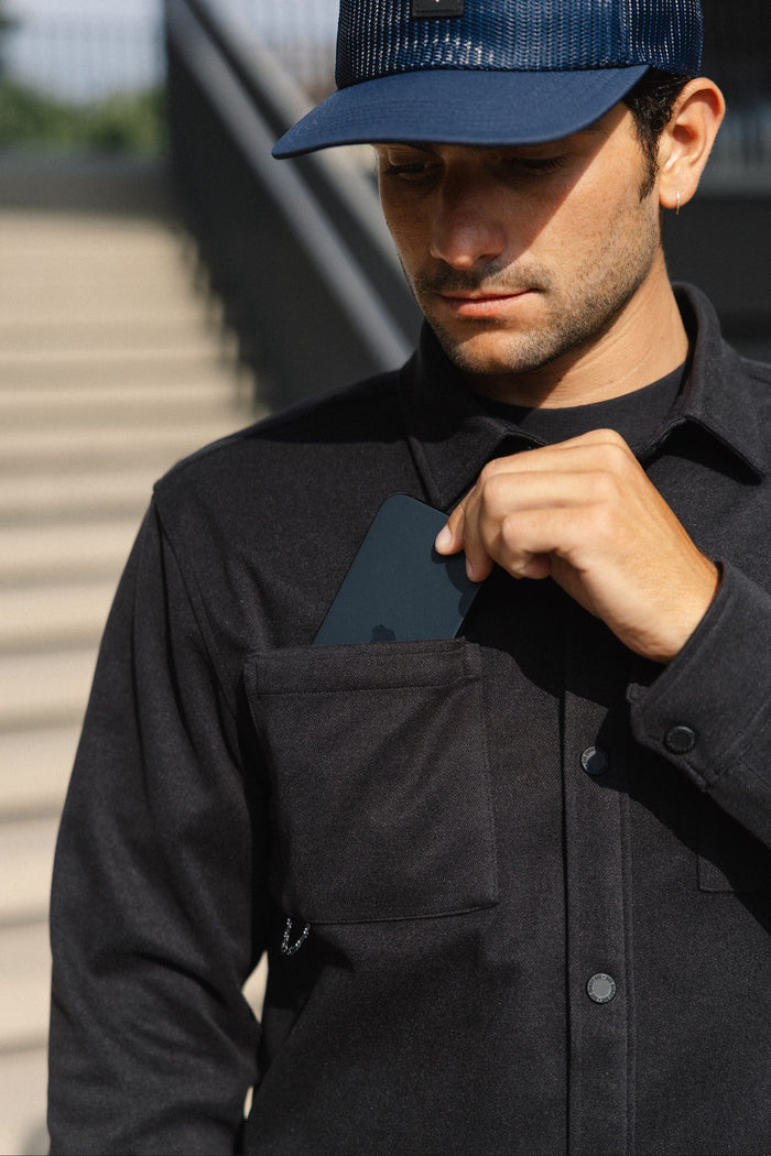 A person wearing the 686 Men's Whisper Knit™ Flannel in black and a blue cap is placing a black smartphone into the front chest pocket of their shirt. A staircase is blurred in the background.