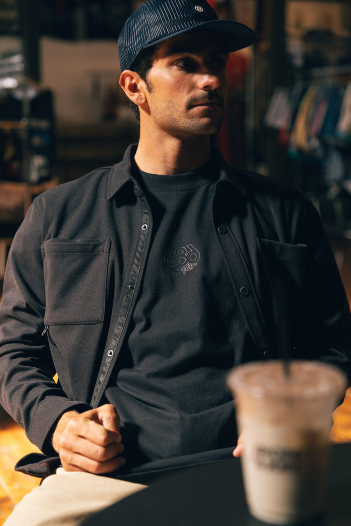 A man wearing the 686 Men's Whisper Knit™ Flannel by 686 and a striped cap sits indoors at a table with a blurry iced coffee in the foreground. Clothing racks are visible in the background, and his stylish polyester flannel adds a touch of modern style to his relaxed look.