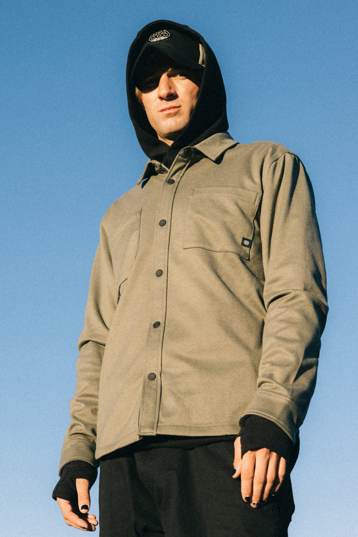 Wearing a khaki button-up jacket over the 686 Men's Whisper Knit™ Flannel by 686 and accessorized with a black beanie, cap, and gloves, a person stands against a clear blue sky looking slightly downward.