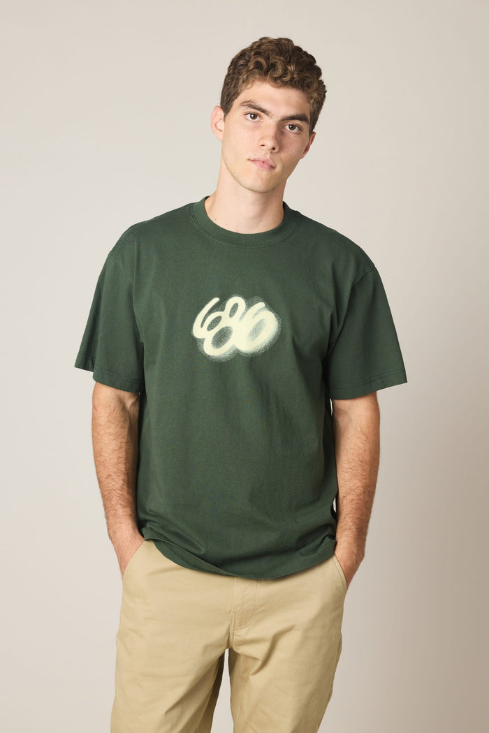 A young man wears the 686 Spray Relaxed SS Tee by 686, a dark green 100% cotton men's T-shirt featuring a spray-painted 686 design, paired with tan pants. He stands against a plain light background, hands in pockets, looking at the camera neutrally.
