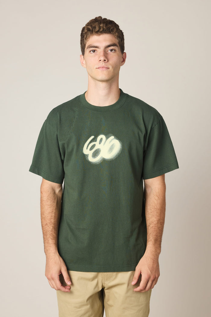A young man with short curly hair wears the 686 Spray Relaxed SS Tee by 686‚Äîa dark green 100% cotton men‚Äôs t-shirt with a white graffiti-style ‚Äú666‚Äù chest design‚Äîand light khaki pants, standing before a plain light background.