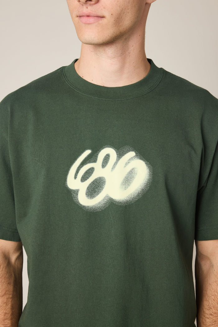 A person wearing the 686 Spray Relaxed SS Tee‚Äîa dark green men‚Äôs t-shirt featuring a white, spray-painted 686 logo. Only the lower face and upper torso are shown, emphasizing its 100% cotton fabric.