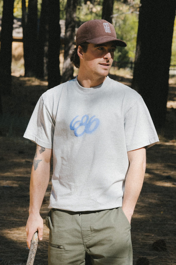 A man in green pants, a brown cap, and the 686 Spray Relaxed SS Tee by 686‚Äîa light gray, 100% cotton men's t-shirt with a blue design‚Äîstands in a wooded area holding a stick and looking to the side.