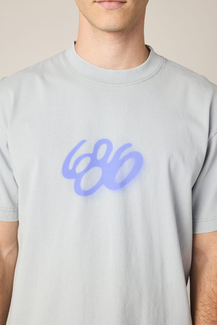 A person wears the 686 Spray Relaxed SS Tee by 686, a light gray men's t-shirt featuring a blue graffiti-style "686" on the chest. Made from soft 100% cotton, it offers comfort; the wearer‚Äôs face is partially out of frame.