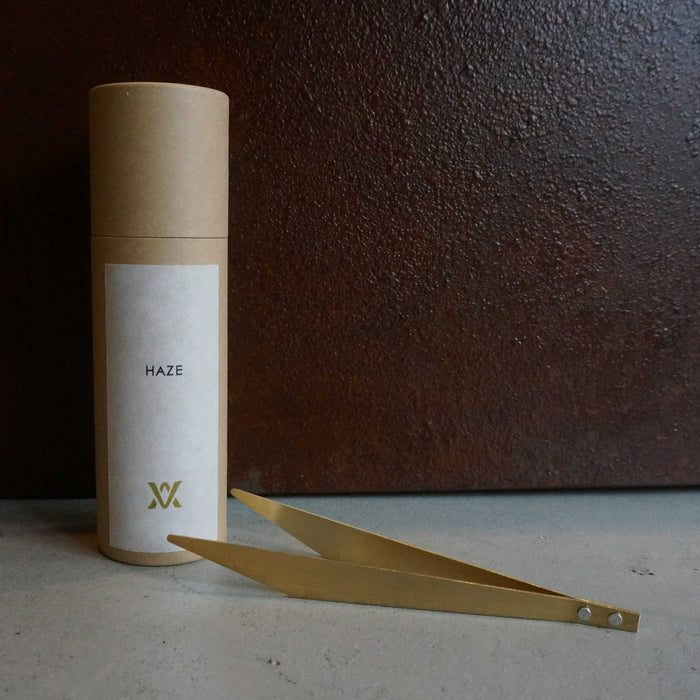 Minimalist brass incense tweezers beside a cylindrical kraft paper tube labeled HAZE, styled on a concrete surface against a dark textured background in a modern product presentation.