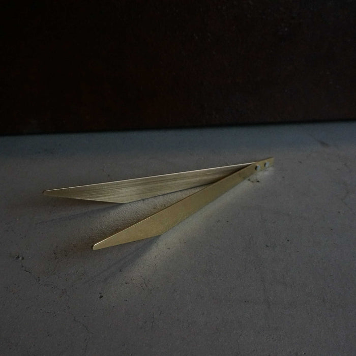 Minimalist brass incense tweezers with a sleek angular design, styled on a concrete surface against a dark moody background in a modern product presentation.