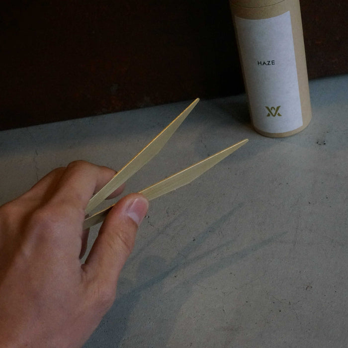Hand holding minimalist brass incense tweezers beside a HAZE kraft paper tube, styled on a concrete surface against a dark textured background in a modern product presentation.