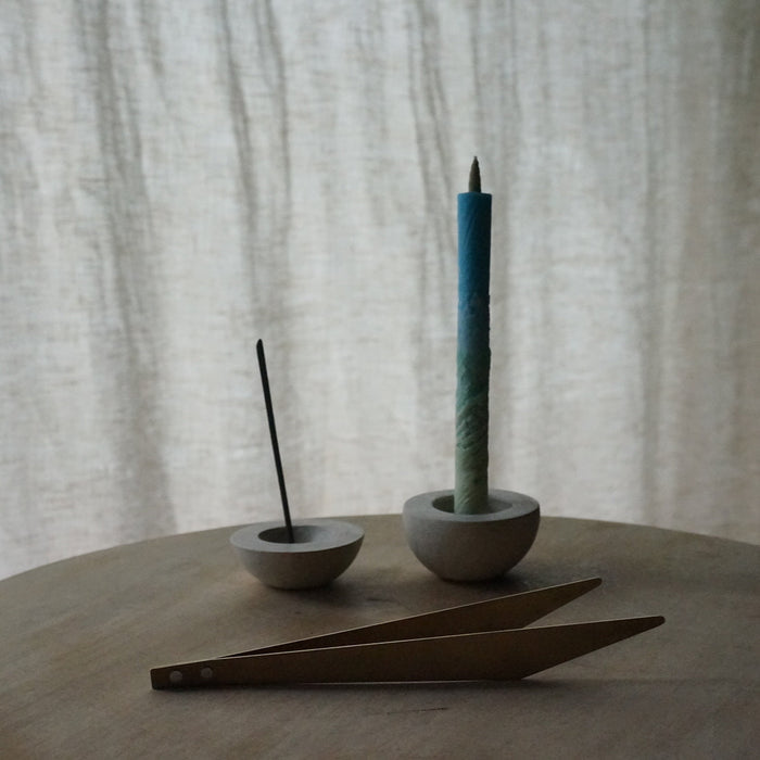 Minimalist brass incense tweezers displayed with a slim incense stick, blue tapered candle, and small stone holders on a round wooden table against soft neutral curtains.