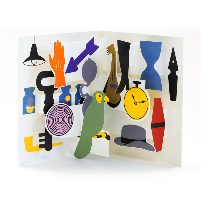 Colorful 3D pop-up greeting card with modern paper illustrations including a green parrot, clock, hat, lamp, perfume bottle, bow tie, and abstract objects in a playful contemporary design.