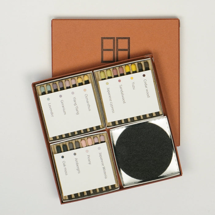 An open square hibi incense match gift set on a white background, showing three compartments of pastel-tipped incense matches labeled with different scents and one compartment containing a round black striker pad, with a brown hibi box lid placed behind the open set.