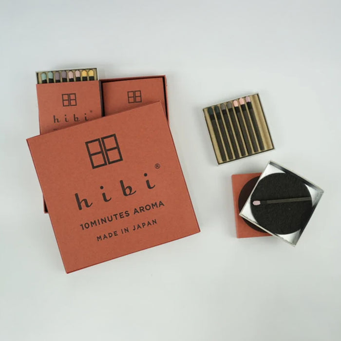 A hibi 10 Minutes Aroma incense match set on a white background, showing brown hibi boxes partially open with pastel-tipped incense matches visible, alongside a separate match tray and a black round striker pad in a small metal holder.