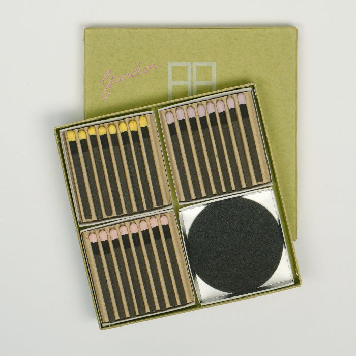 An open square hibi incense match gift set on a white background, showing three compartments of pastel-tipped incense matches and one compartment with a round black striker pad, with a green hibi box lid placed behind the open set.