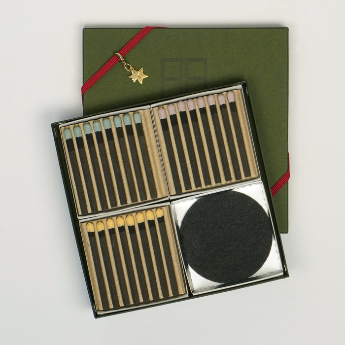 An open square matchbox gift set displayed on a white background, showing neatly arranged matches with pastel-colored tips in four compartments, alongside a round black striker pad. A green cover tied with a red ribbon and a small gold star charm rests beneath the open box.