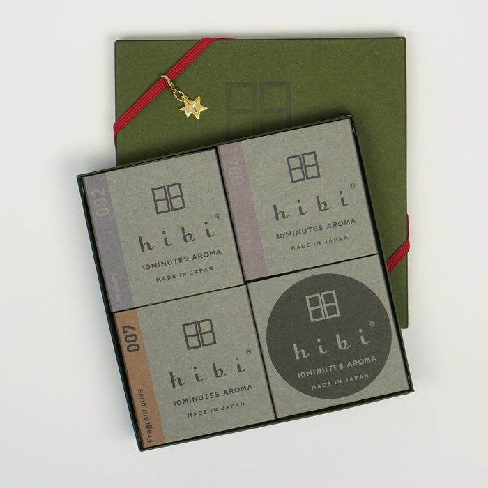 An open square hibi gift box displayed on a white background, showing four minimalist gray packages labeled “hibi 10 Minutes Aroma,” arranged in a grid. A green cover tied with a red ribbon and a small gold star charm sits beneath the open box, creating a refined, gift-ready presentation.