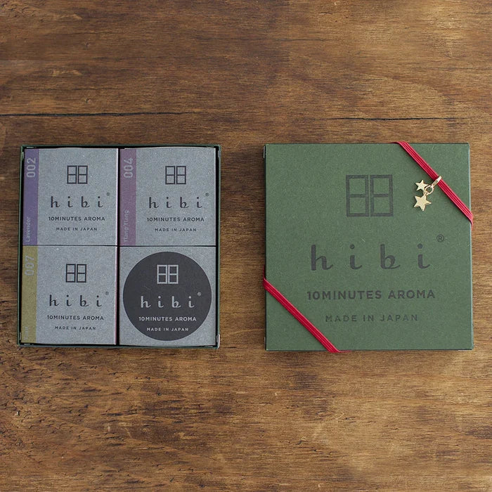 A hibi 10 Minutes Aroma gift set displayed on a wooden surface, showing an open box with four minimalist gray incense match packages on the left and a closed green gift box on the right, wrapped with a red ribbon and finished with a small gold star charm.