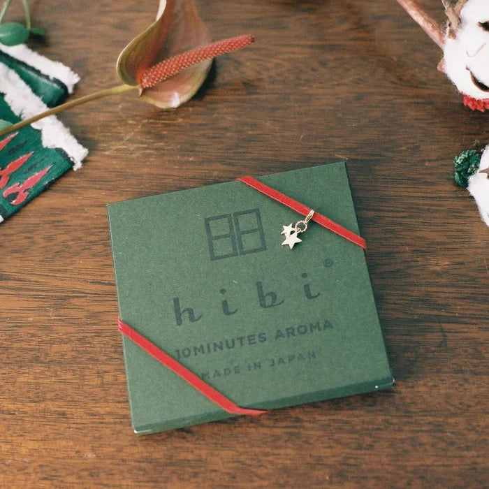 A closed hibi 10 Minutes Aroma gift box resting on a wooden surface, wrapped with a red ribbon and accented by a small gold star charm, surrounded by festive décor elements for a holiday-inspired presentation.