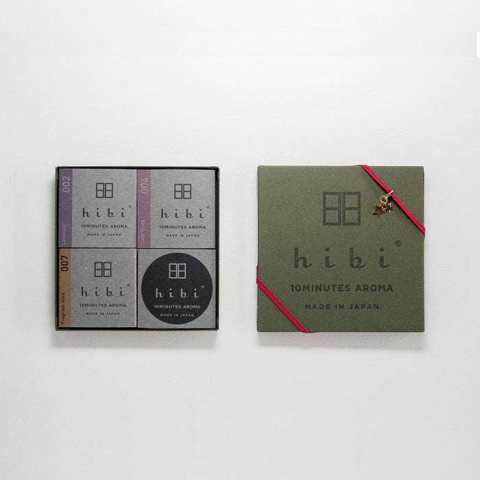A hibi 10 Minutes Aroma gift set on a white background, with an open box on the left showing four gray incense match packages arranged in a grid, and a closed green box on the right wrapped with a red ribbon and finished with a small gold star charm.